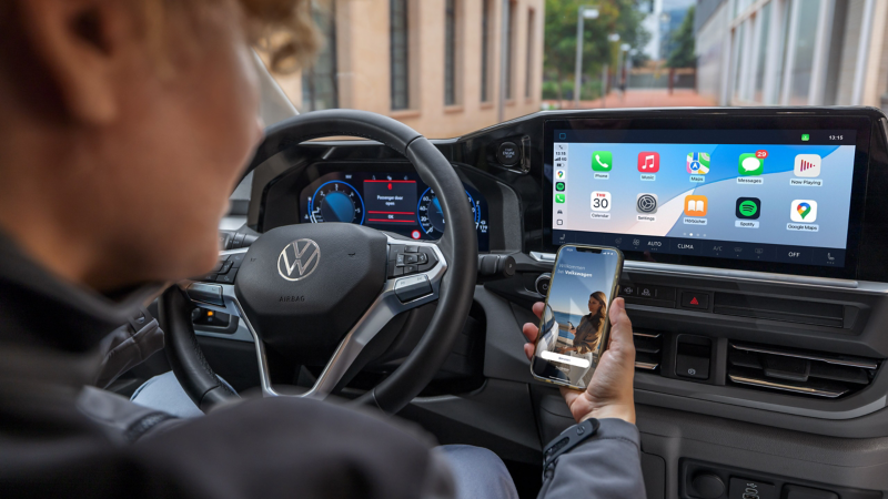 Eine Person am Steuer eines parkenden VW Transporter blickt auf ein Smartphone mit geöffneter VW Connect App.