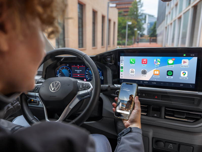 Eine Person am Steuer eines parkenden VW Transporter blickt auf ein Smartphone mit geöffneter VW Connect App.