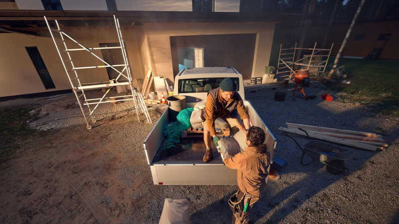 Zwei Bauarbeiter in Arbeitskleidung entladen einen VW Transporter Pritschenwagen, der auf einem Schotter- und Sandgrund vor einer Baustelle steht.