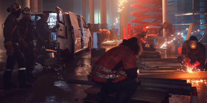 Mehrere Bauarbeiter in Schutzkleidung arbeiten in einer dunklen, feuchten Industriehalle, wobei Funken fliegen, während im linken Bildhintergrund ein weißer VW Transporter mit geöffneten Türen steht.