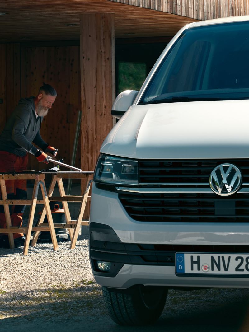 Ein Volkswagen Transporter 6.1 Kastenwagen vor einer Holzwerkstatt. Im Hintergrund arbeitet ein Schreiner.