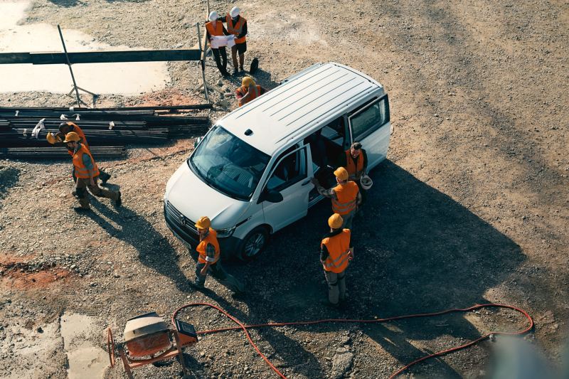Ein VW Transporter auf einer Baustelle. Einige Bauarbeiter um das Fahrzeug rum.