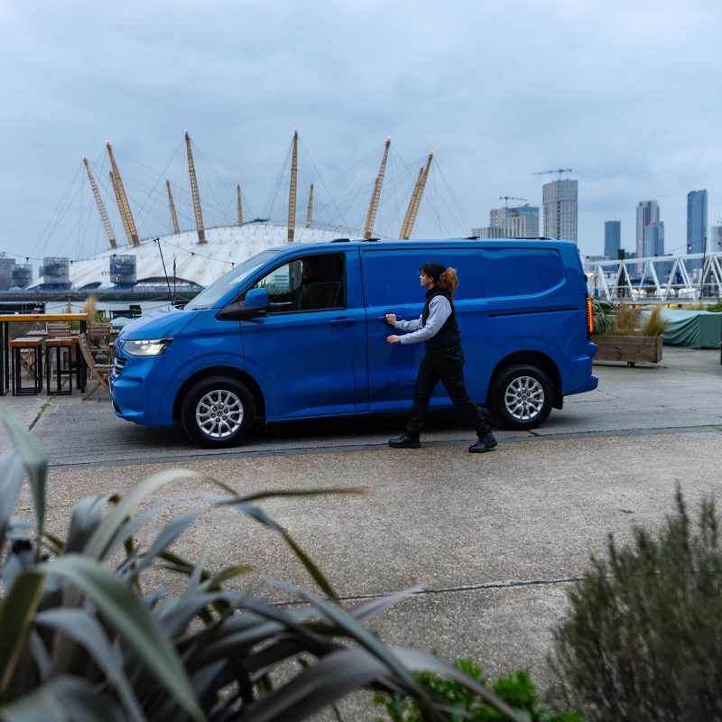 A person approaches a blue Transporter Panel van parked near the waterfront, with the O2 Arena and urban skyline in the background under a cloudy sky.
