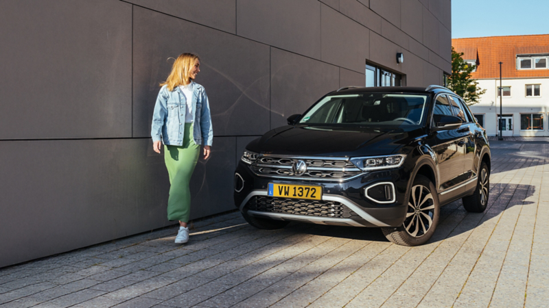 Un T-Roc noir est garé devant un bâtiment, tandis qu'une femme passe devant.