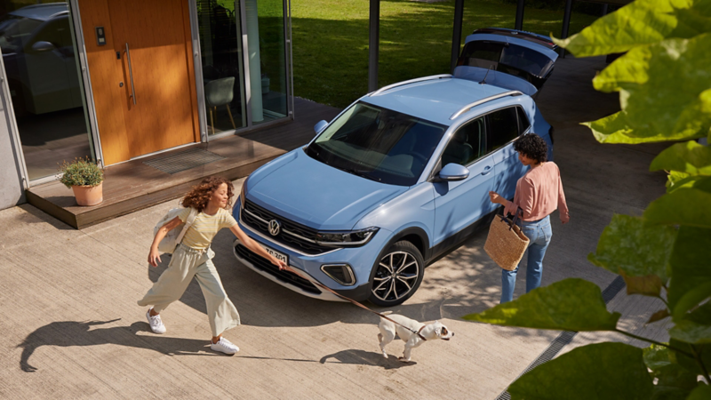 Ein blauer VW T-Roc Cabriolet steht vor einem Haus. Daneben stehen eine Frau und ein Mädchen mit einem Hund an der Leine.