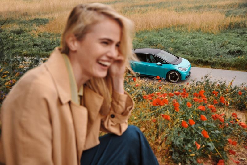 Volkswagen ID.3 en una carretera detrás de una chica sonriendo sentada en un campo de flores 