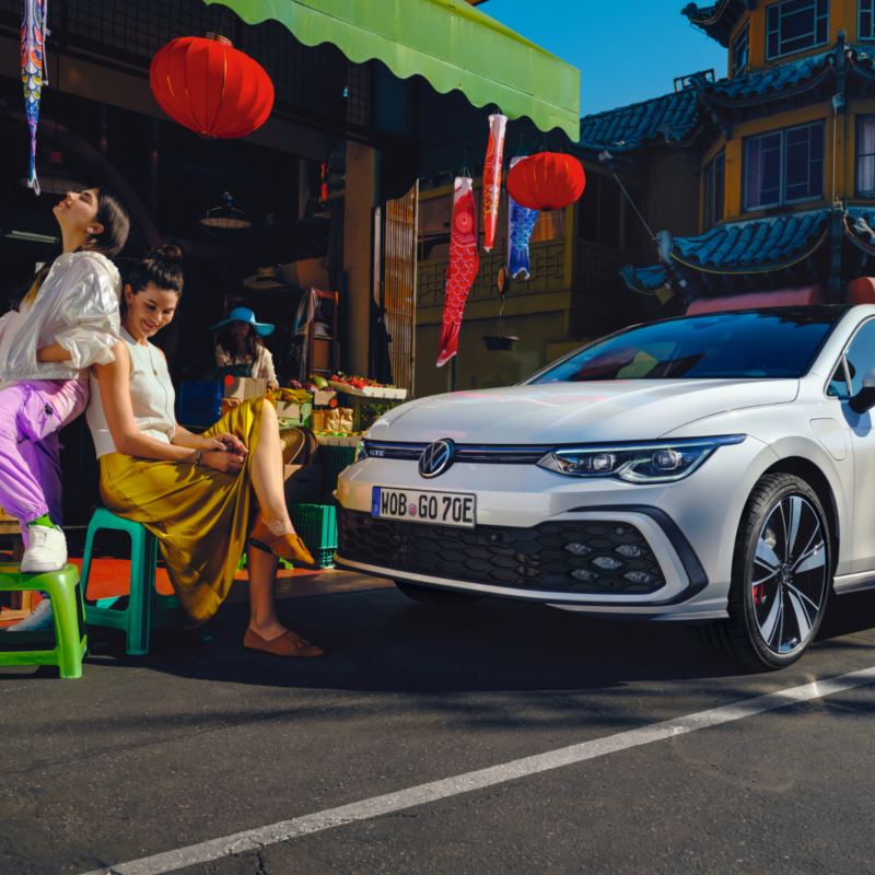 Dos chicas y un Golf híbrido enchufable blanco frente a un puesto en un mercado chino