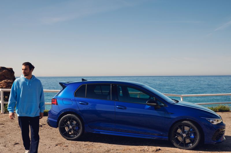 Un chico junto a un Volkswagen Golf R azul visto de costado y aparcado junto al mar