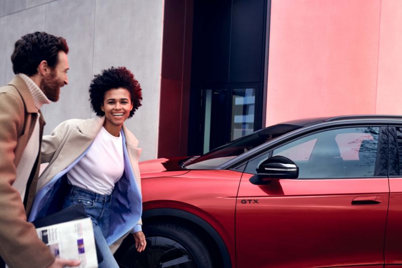 Volkswagen ID.4 GTX en rojo aparcado al borde de la carretera, vista lateral. Una mujer y un hombre pasan por delante del vehículo, sonriendo.