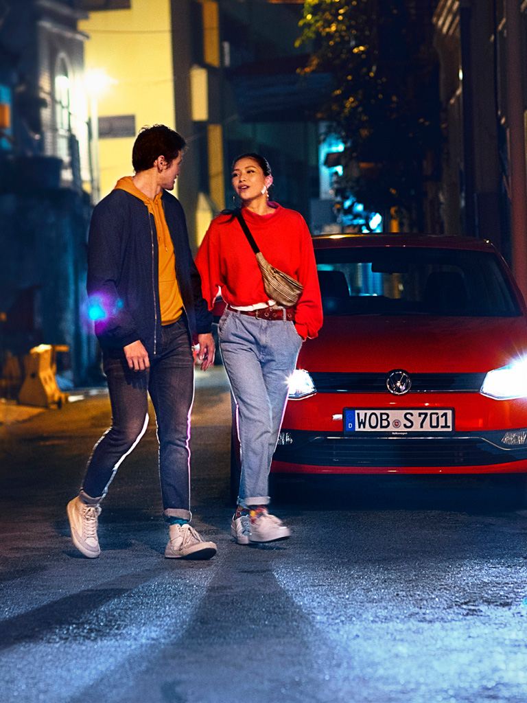 Una pareja de la mano caminando de noche delante de un Volkswagen rojo con los faros encendidos