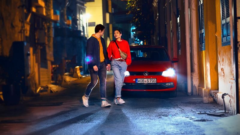 Una pareja de la mano caminando de noche delante de un Volkswagen rojo con los faros encendidos