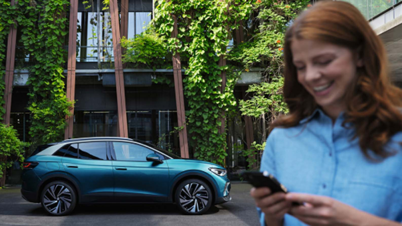Un plano lateral de un Volkwagen eléctrico azul estacionado en un patio de una casa con vegetación y en el lado derecho en primer plano desenfocado una persona contenta con su teléfono móvil