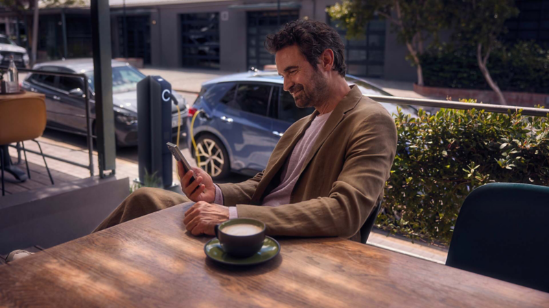 Una persona revisando su movil con un café junto a un Volkswagen conectado a carga