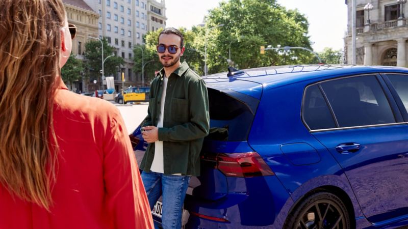 Un chicho apoyado en un Volkswagen Golf 8 R azul hablando con una chica en la calle