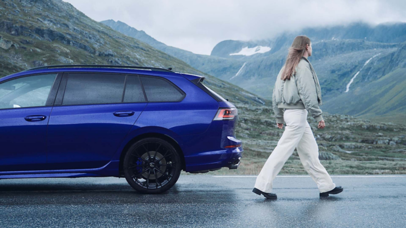 Una mujer pasando por detrás de un Volkswagen Golf 8 Variant R azul parado frente unas montañas