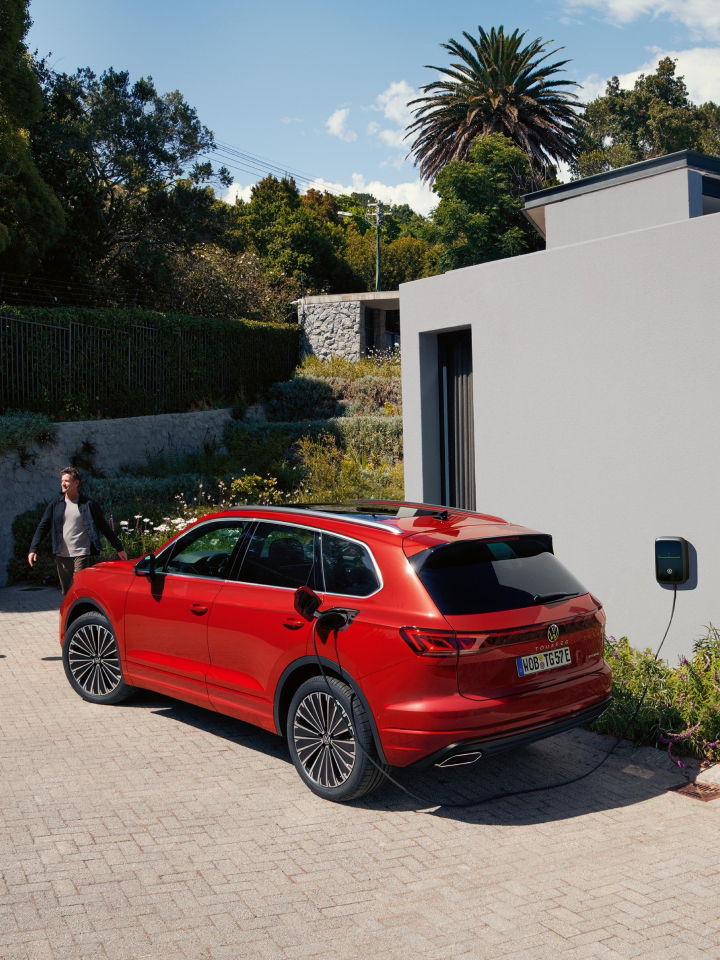 Un hombre al lado de un Volkswagen de color rojo conectado a carga estacionado fuera de una casa