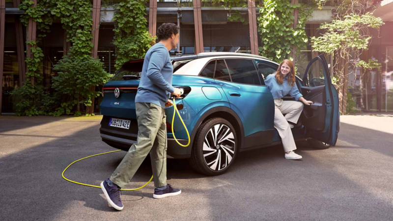 Un hombre acercándose con un cable cargador de un Volkswagen ID4 azul con una mujer asomando desde el asiento del copiloto