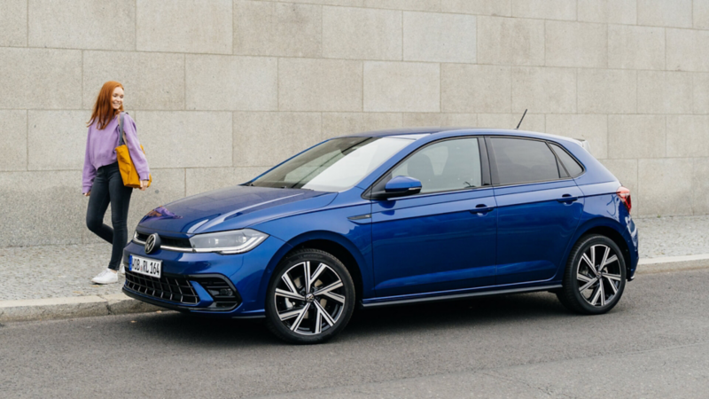 Una chica al lado de un Volkswagen Polo de color azul oscuro estacionado en la calle