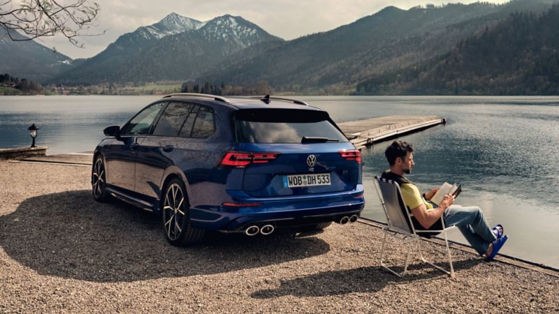 chico leyendo al lado de un Volkswagen Variant azul oscuro estacionado junto a un lago