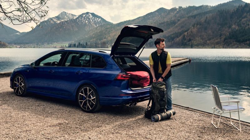 hombre junto a un Volkswagen Golf Variant azul con el maletero abierto estacionado junto a un lago