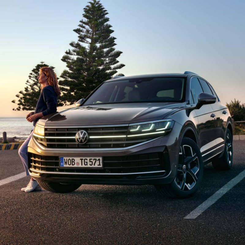 Una chica apoyada en un Volkswagen Touareg visto de frente al atardecer