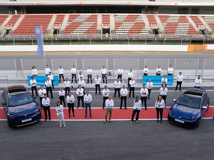 Foto picada en plano abierto con el estadio, dos coches Volkswagen a los laterales y profesionales de la conducción posando