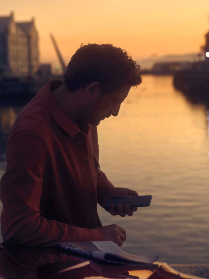 Hombre mirando su móvil delante del mar, apoyado en un Arteon Shooting Brake rojo