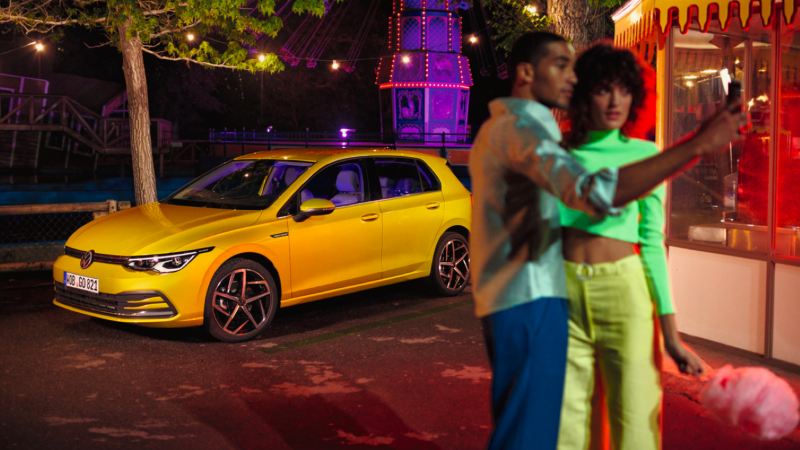 Una pareja haciéndose un selfie en una feria con unVolkswagen Golf 8 amarillo detrás