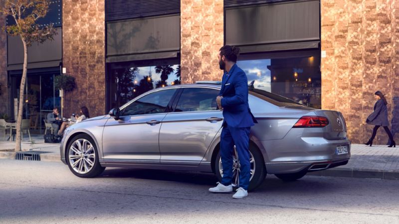 Hombre de azul de pie al costado de un Volkswagen Passat gris aparcado en la calle
