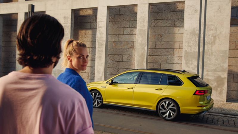 Chica mirando a la cámara detrás de un chico y Volkswagen amarillo aparcado al fondo