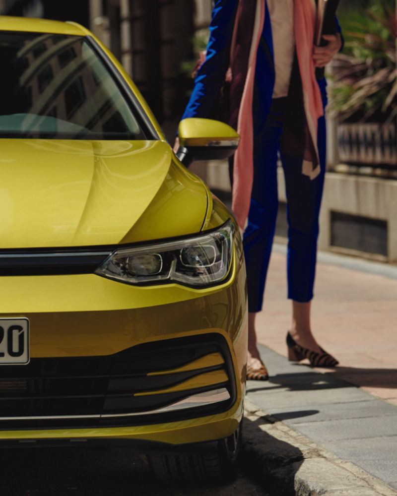 Detalle del frontal de un Volkswagen Golf 8 amarillo lima aparcado con una mujer detrás