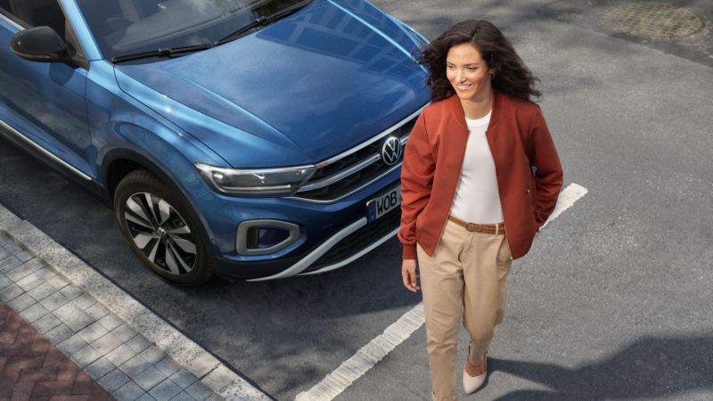 Una chica al lado de un Volkswagen de color azul estacionado en una plaza de parking