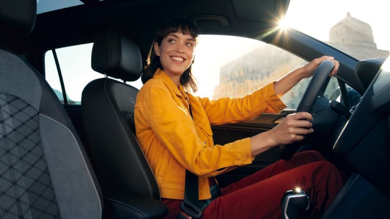 Chica joven sonriendo vestida de amarillo sentada al volante de un Volkswagen