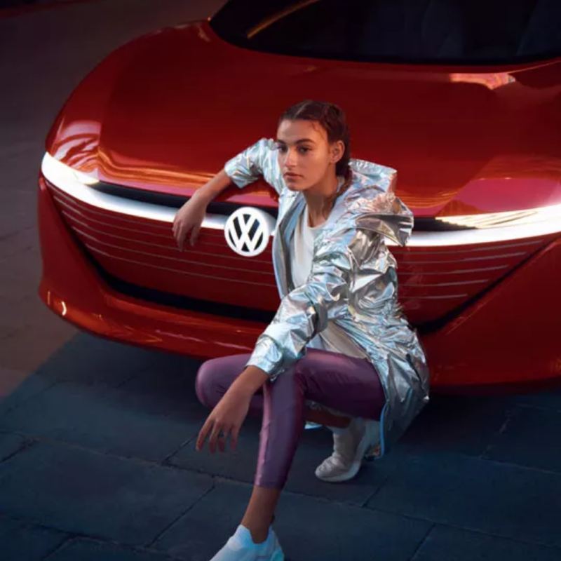Chica posando de cuclillas apoyada en el frontal de un Volkswagen ID. Vizzion rojo