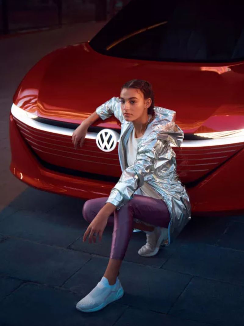Chica posando de cuclillas apoyada en el frontal de un Volkswagen ID. Vizzion rojo
