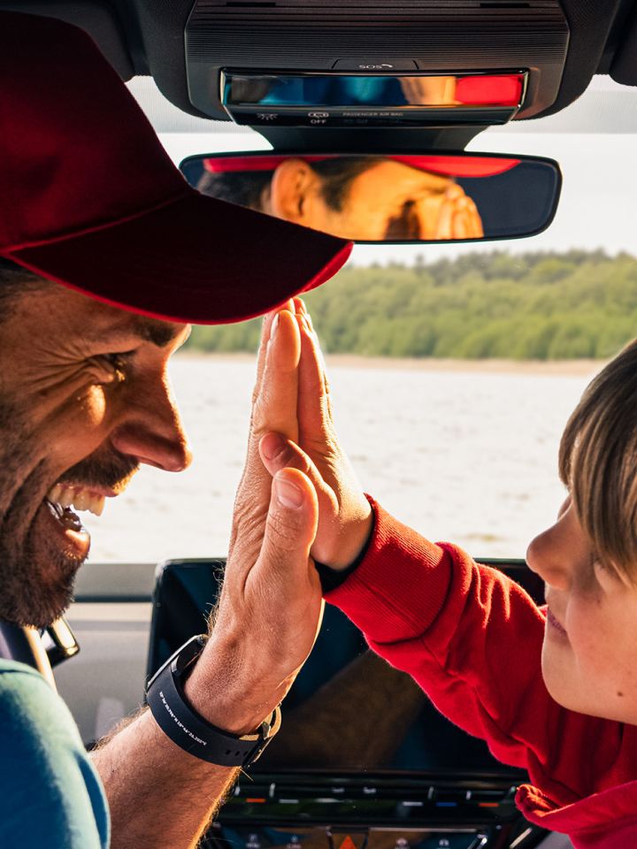 Un padre con su hijo sonriendo y chocando las manos dentro de un Volkswagen y un lago al fondo