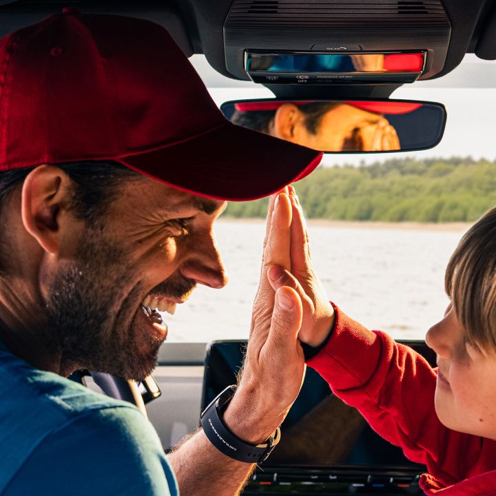 Un padre con su hijo sonriendo y chocando las manos dentro de un Volkswagen y un lago al fondo