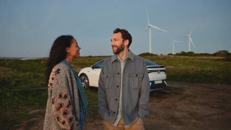 Pareja de pie delante de un vehículo eléctrico de Volkswagen con energía eólica de fondo