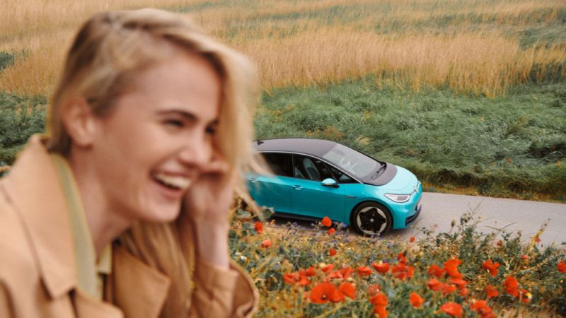 Chica hablando por móvil en primer plano con un Volkswagen ID.3 de fondo en una carretera de campo