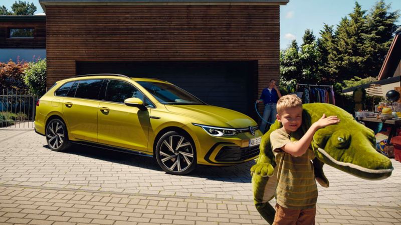 Una un niño con un peluche frente a un Volkswagen Golf Variant amarillo aparcado en casa, accesorios Volkswagen