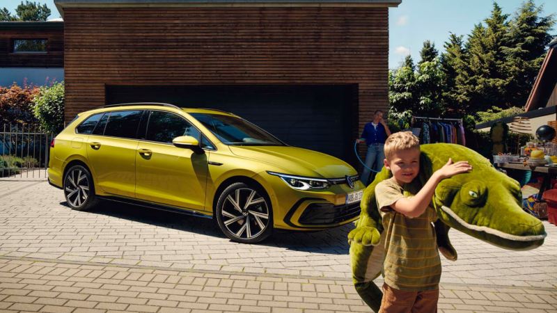 Un niño con un peluche delante de un Volkswagen Golf 8 Variant amarillo aparcado en casa