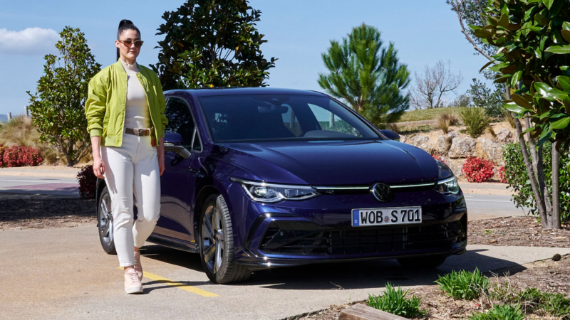 Una chica de pie junto a un Volkswagen Golf azul aparcado, accesorios Volkswagen