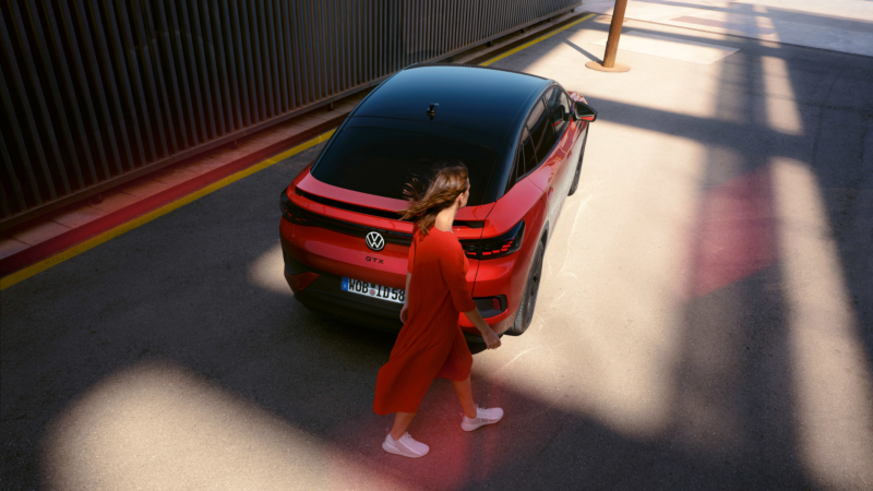 Una chica vestida de rojo pasando detrás de un Volkswagen ID.5 GTX rojo parado en un hangar