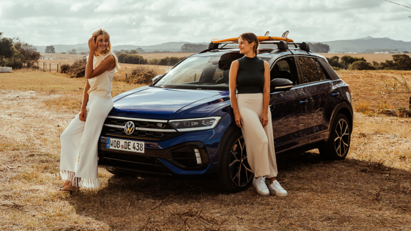 Dos chicas apoyadas en la parte delantera de un Volkswagen T-Roc R azul estacionado en el campo