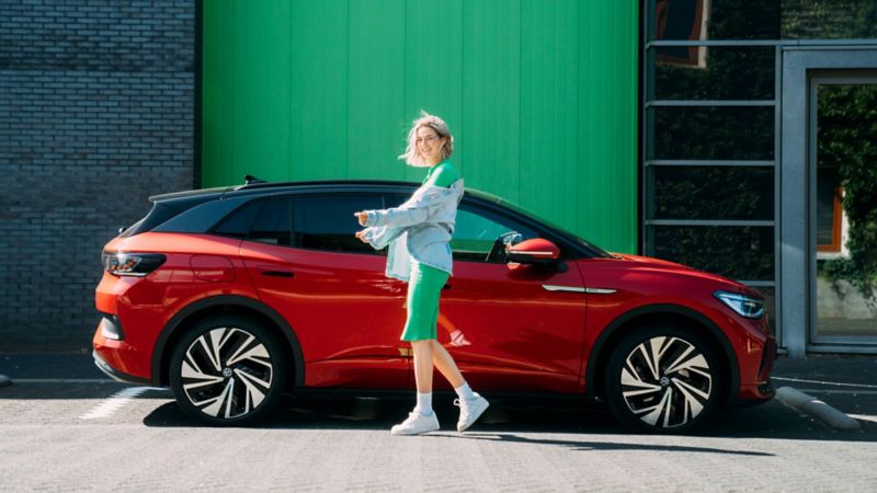 Chica sonriendo de pie frente a un Volkswagen ID.4 rojo visto de costado y un muro verde