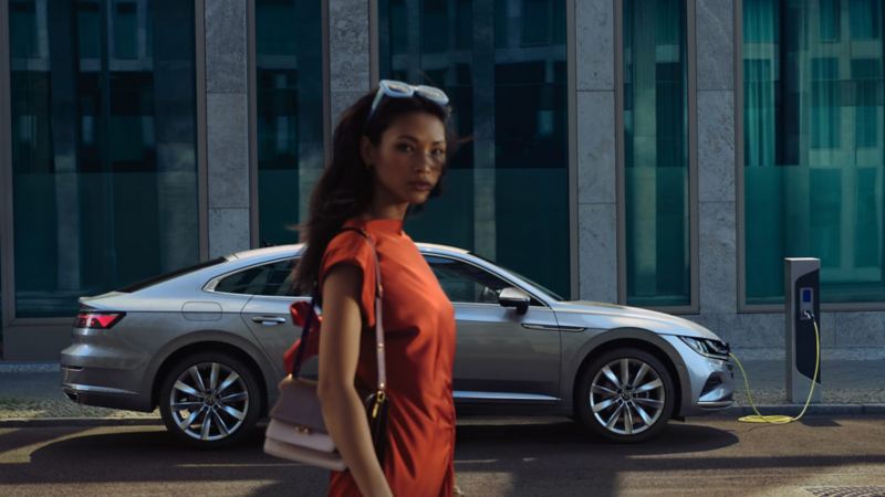 Una mujer caminando delante de un Volskwagen Arteon hibrido enchufable conectado a una estación de carga