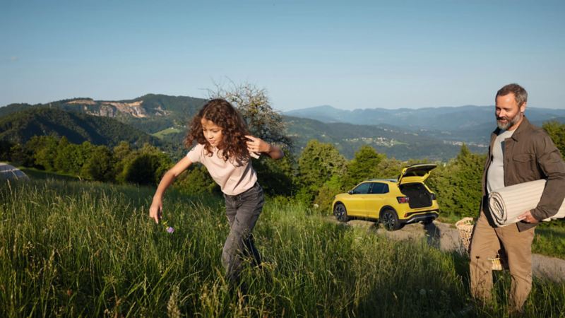 Una niña corre por el campo con su padre detrás, y su Volkswagen amarillo de fondo