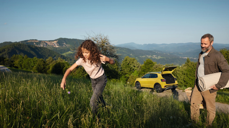 Una niña corre por el campo con su padre detrás, y su Volkswagen amarillo de fondo