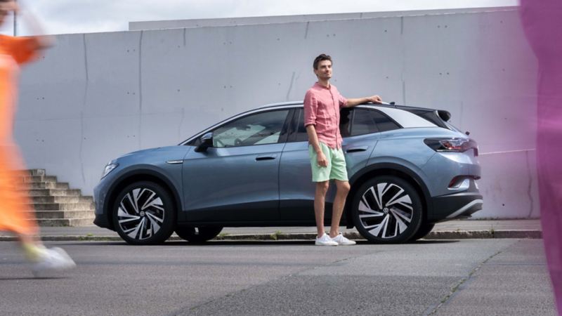 Chico de pie junto a un Volkswagen ID.4 gris aparcado frente a un muro de concreto