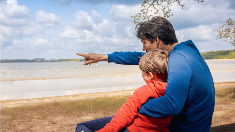 Un padre indicando el horizonte junto a su hijo sentados en la orilla de un lago